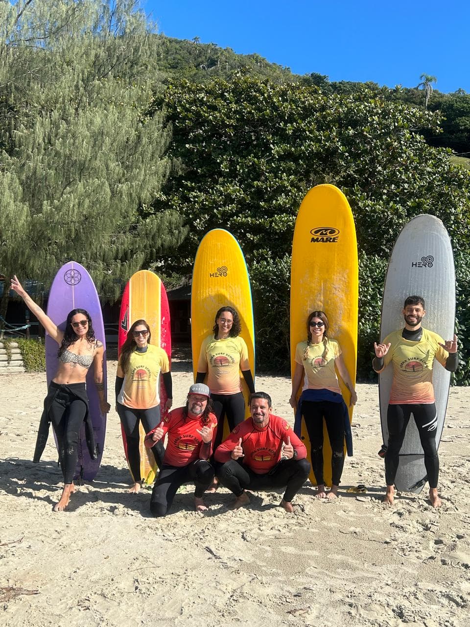 Grupo de alunos e instrutores com pranchas de surf em frente à vegetação da praia