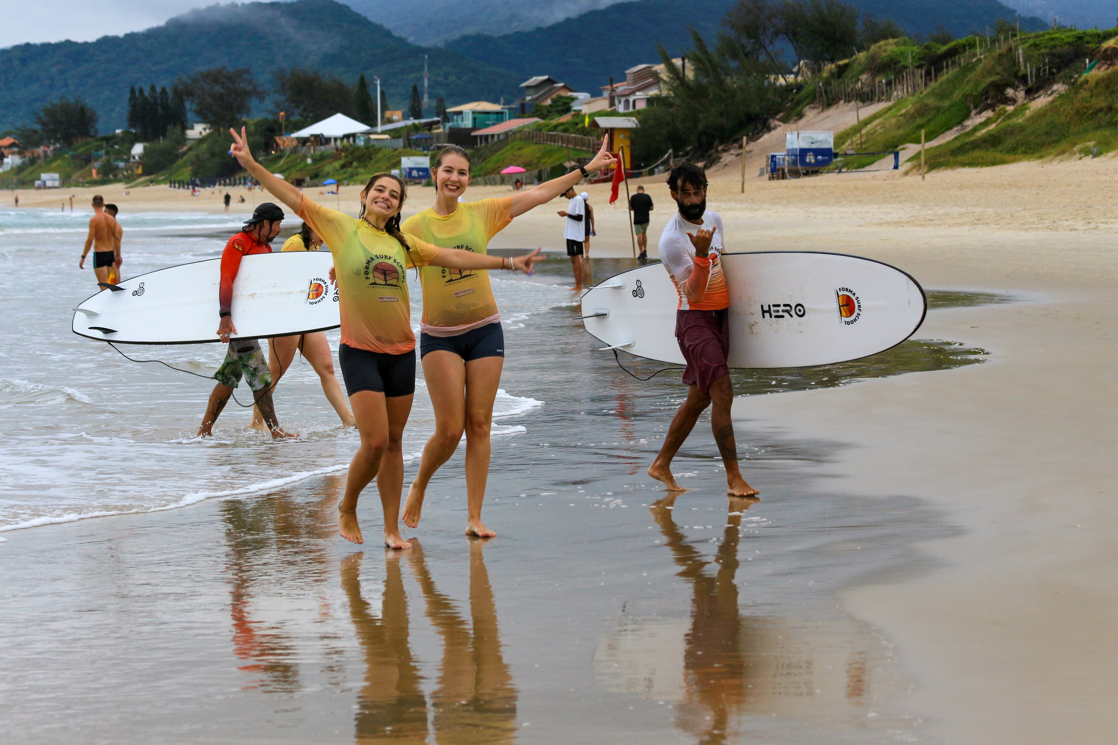 Alunas comemorando ao caminhar na praia com as pranchas após a aula de surf