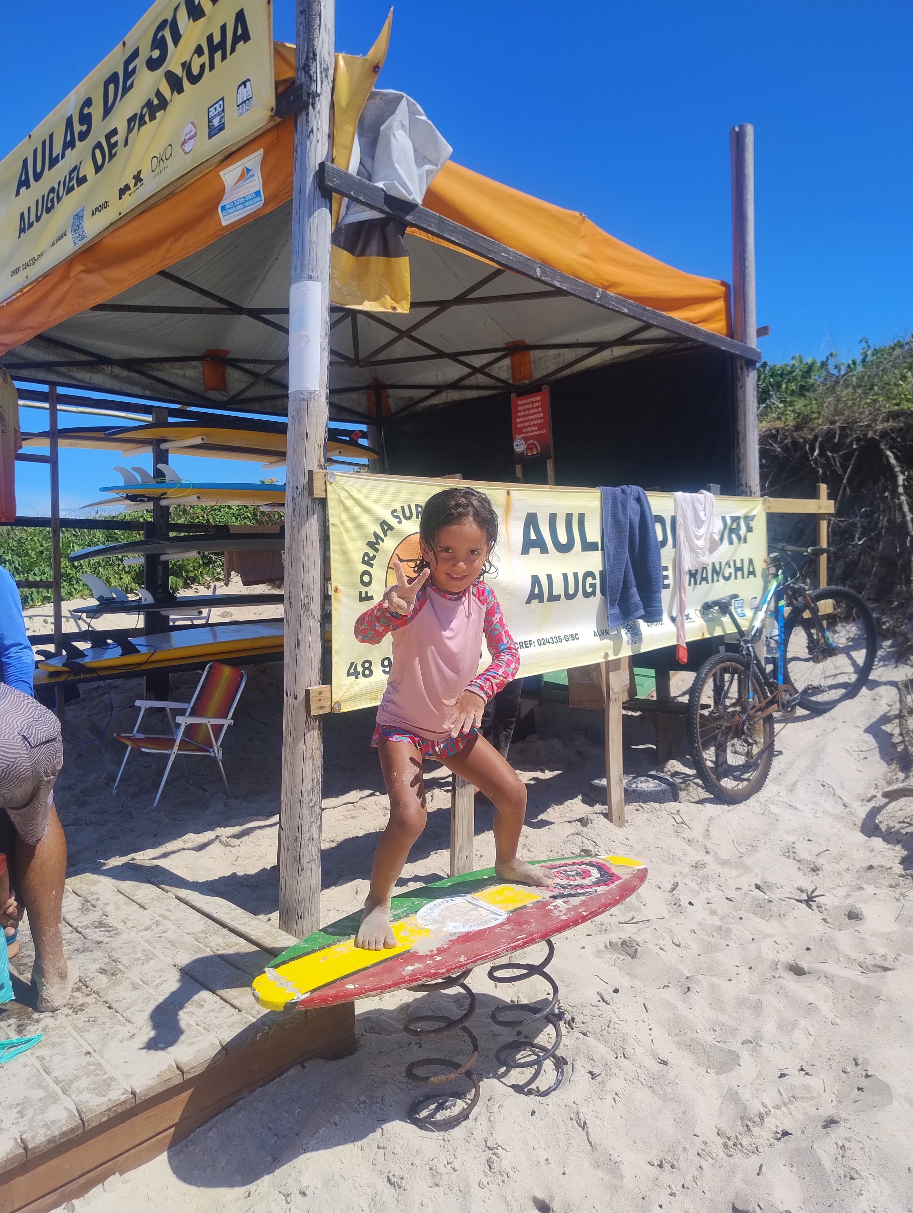 Criança sorridente sobre uma prancha de surf em frente à barraca da escolinha